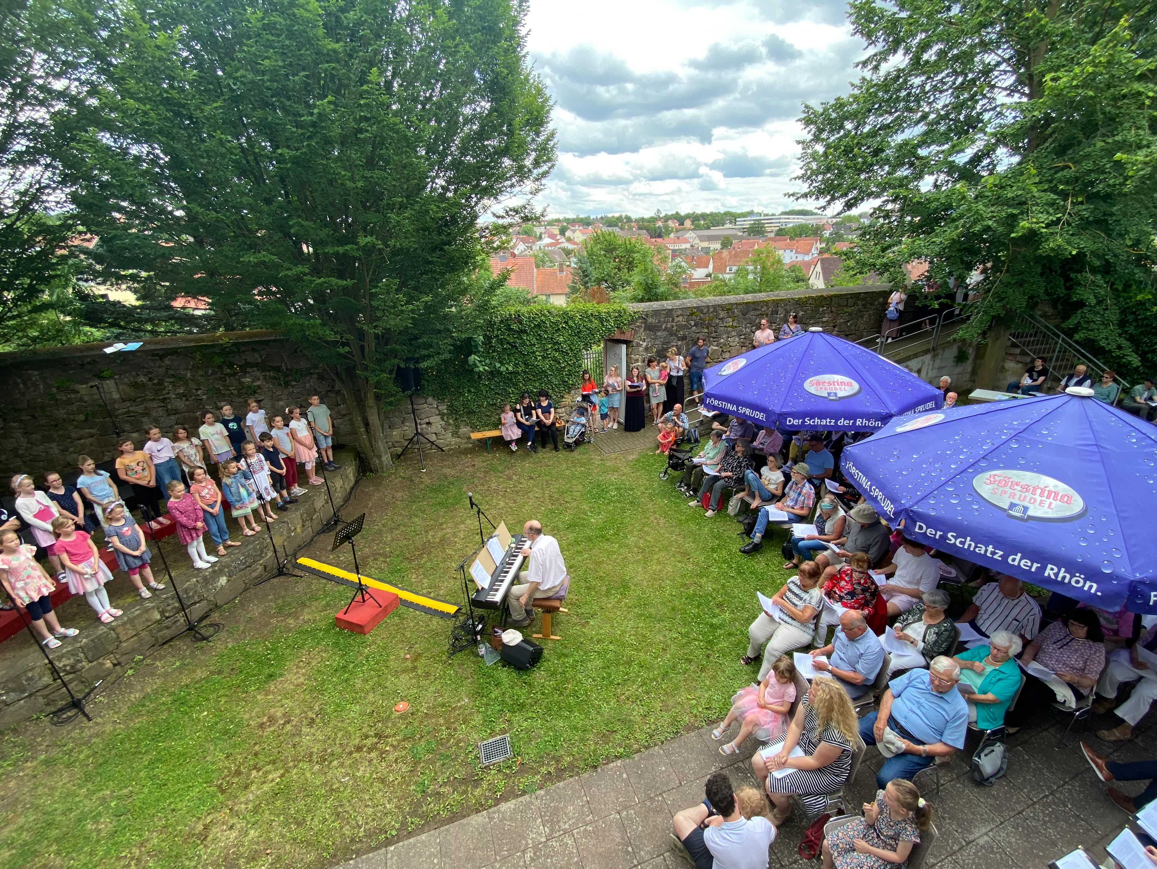 Volksliedersingen. Personen auf einer Wiese, die einem Chor zuhören. 